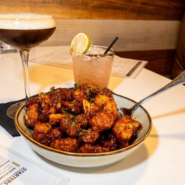 A bowl of saucy cauliflower bites with two cocktails and a menu on a white table.