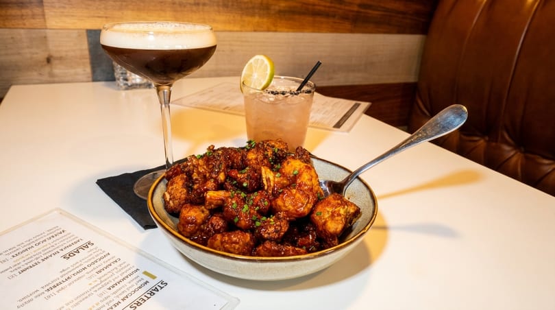 A bowl of crispy cauliflower with two cocktails and a menu on a white table.