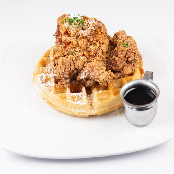 Fried chicken and waffles with powdered sugar, chives, and a side of syrup on a white plate.