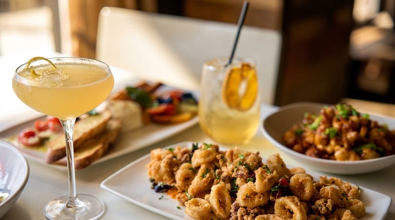 A table filled with various dishes, including fried calamari, pasta, and two cocktails.
