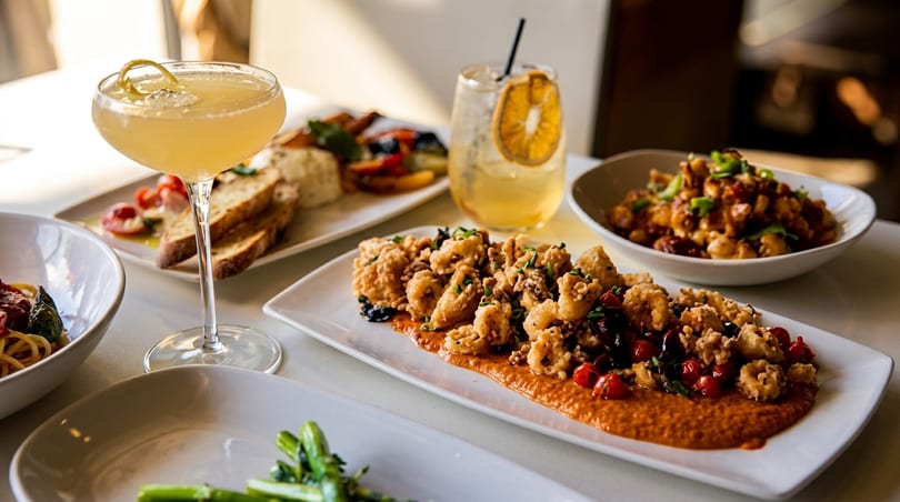 A table laden with various dishes including fried calamari, pasta, and cocktails.