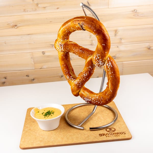 Large salted pretzel on a metal stand with a bowl of cheese dip and chives on a Brauhaus Pretzel cutting board.