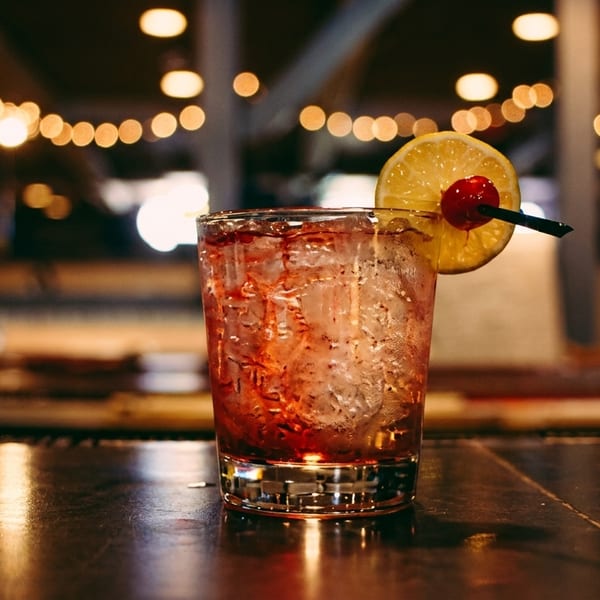 A red cocktail with ice, garnished with a lemon slice and cherry, on a dark bar.