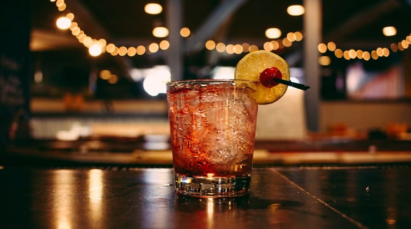 A red cocktail with ice, garnished with a lemon slice and cherry, on a dark bar.