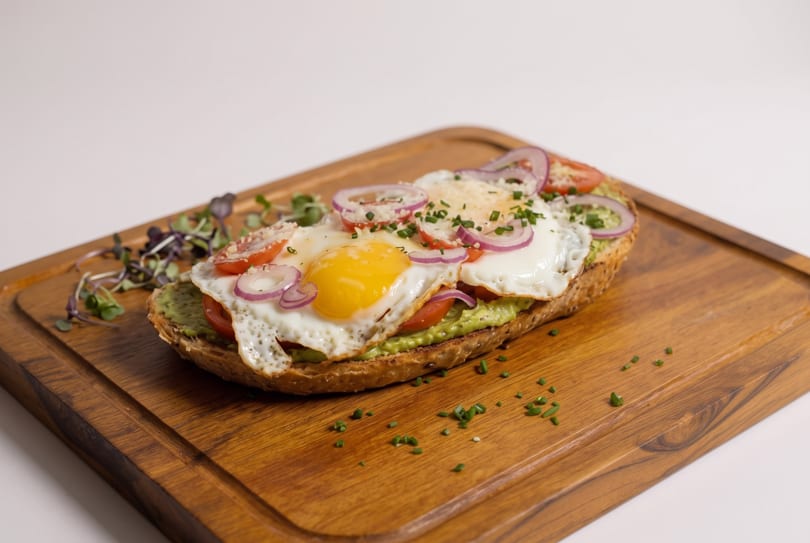 Avocado toast with fried eggs, sliced tomatoes, red onions, and chives on a wooden board.