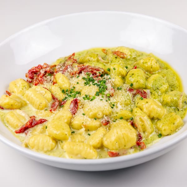 Gnocchi in creamy pesto sauce with sun-dried tomatoes, parmesan, and chives in a white bowl.