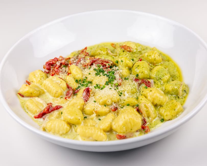 Gnocchi in creamy pesto sauce with sun-dried tomatoes, parmesan, and chives in a white bowl.