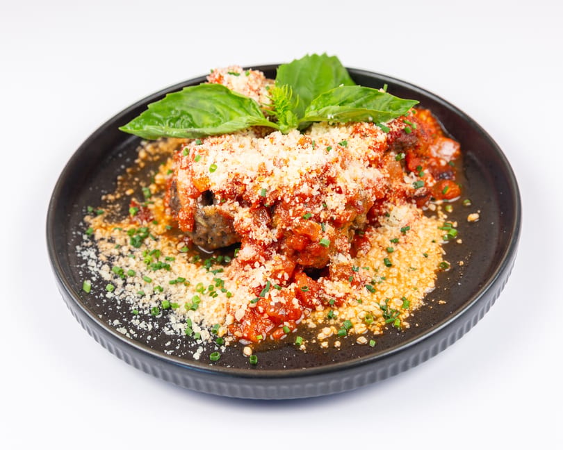 Meatballs covered in red sauce, grated cheese, basil, and chives on a dark plate.