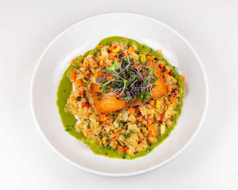 Crispy salmon fillet on vegetable quinoa with vibrant green sauce and fresh microgreens.