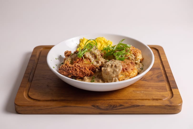 Fried chicken and biscuits with gravy, scrambled eggs, and green onions in a white bowl on a wooden board.