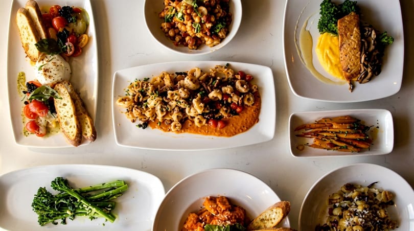 Overhead view of various Italian dishes, including pasta, burrata, salmon, and vegetables on a white table.