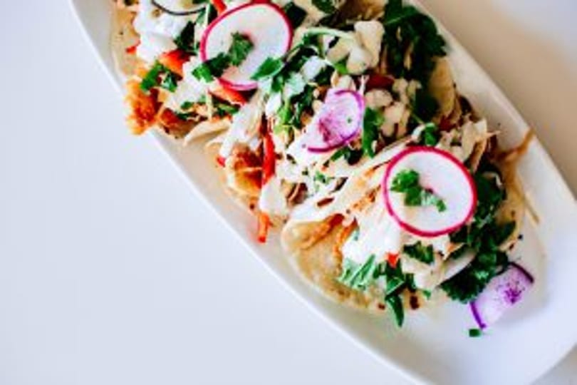 Three vibrant tacos with radish, cabbage, and cilantro on a white platter.