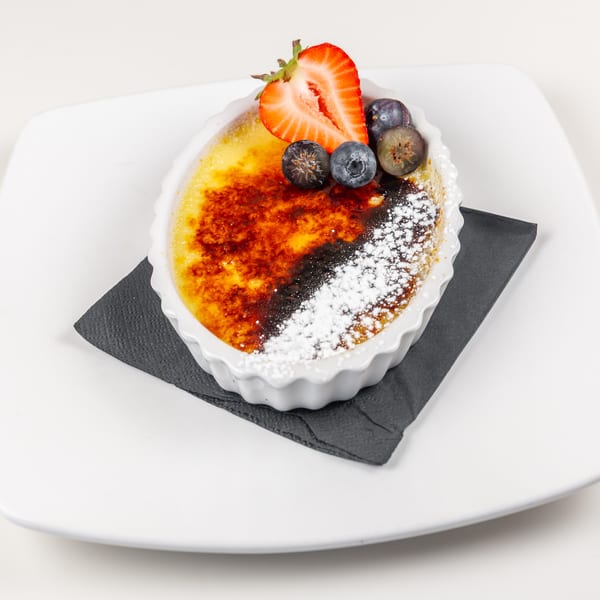 Creme brulee with a caramelized sugar topping, powdered sugar, strawberry, and blueberries on a white plate.