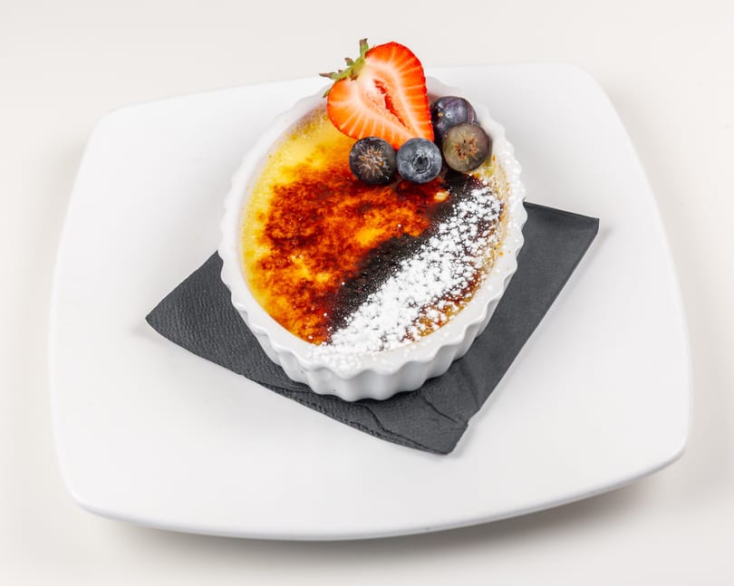Creme brulee with a caramelized sugar topping, powdered sugar, strawberry, and blueberries on a white plate.