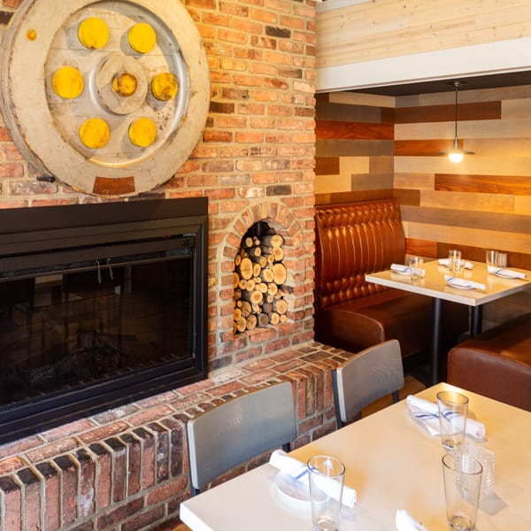 Cozy restaurant interior featuring a brick fireplace, decorative wheel, stacked wood, and set tables with brown booths.