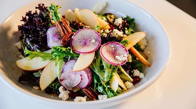 A vibrant salad with mixed greens, sliced apples, radishes, pecans, and crumbled cheese in a white bowl.