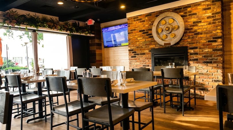 Restaurant interior with wooden tables and chairs, brick wall, fireplace, TV, and large windows.